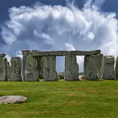 Antico cromlech di pietre imponenti su un prato verde, sotto un cielo drammatico con nuvole bianche e blu.