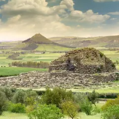 Antico nuraghe in pietra, con scale esterne, immerso in un paesaggio verdeggiante con colline ondulate e un cielo nuvoloso.
