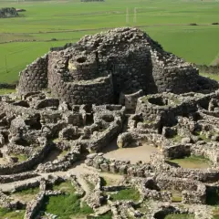 Antica fortezza nuragica in pietra, con torri e mura a vista, immersa in un paesaggio rurale verdeggiante. I nuraghi dove si trovano sono testimoni di un passato millenario.