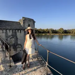 Donna con cappello e giacca in pelle, in piedi sul ponte di pietra vicino al fiume, con un edificio antico sullo sfondo, che ricorda un altare rupestre di Santo Stefano.