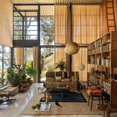 Interno della Eames House con grandi finestre, mobili iconici, piante rigogliose e una libreria piena.