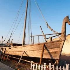 Una nave fenicia in legno, con prua a forma di testa di cavallo, è esposta su un terreno sabbioso vicino al mare.