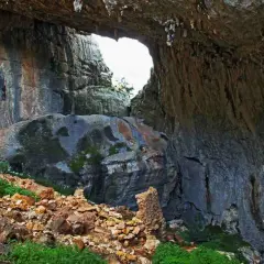 Ingresso di una grotta con rocce stratificate e vegetazione. Un arco naturale si apre verso l'esterno, rivelando un paesaggio luminoso. Un sito archeologico suggestivo.
