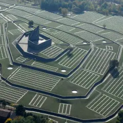 Vista aerea del cimitero tedesco al Passo della Futa, con file ordinate di croci bianche su prati verdi e un imponente monumento centrale.
