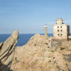 Il faro della Maddalena, con la sua torre e l'edificio adiacente in rovina, si erge su una costa rocciosa battuta dal mare.