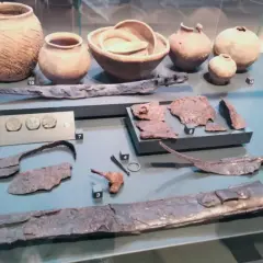 Reperti archeologici, tra cui vasi, monete e armi, simili a quelli della patera di Parabiago, esposti in un museo.