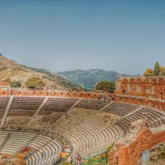 Antico teatro greco in Italia, con gradinate in pietra e rovine in mattoni rossi, incastonato tra colline verdi e un cielo azzurro.