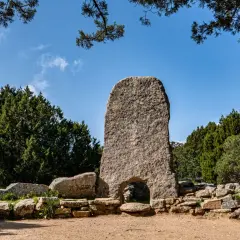 Un imponente menhir con un arco alla base, circondato da pietre antiche e vegetazione rigogliosa. Tra le tombe dei giganti più belle, questo sito archeologico incanta.