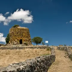 Antichi nuraghi sardi, testimoni di un passato di guerra e civiltà. Un sentiero di pietra conduce verso la torre, immersa in un paesaggio arido sotto un cielo azzurro.