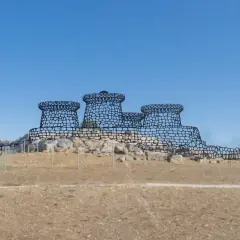 Disegno stilizzato del nuraghe Genna Maria, con torri tronche e mura merlate, su uno sfondo di cielo azzurro e terreno brullo.