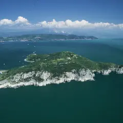 Isola verdeggiante con scogliere bianche, mare blu intenso e nuvole bianche. Si intravede la **grotta dei colombi** tra le rocce.