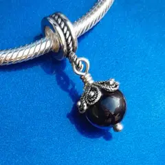 Un ciondolo con una pietra nera lucida, ispirato agli antichi amuleti sardi, pende da un bracciale d'argento su sfondo blu.
