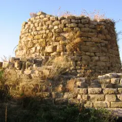 Il nuraghe Piscu, con le sue pietre antiche e l'erba secca, si staglia contro il cielo azzurro, testimone silenzioso di storie millenarie.