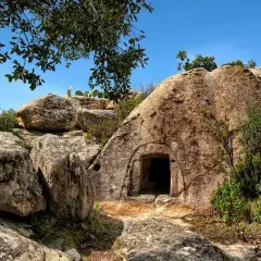 Ingresso di una delle **domus de janas**, tombe ipogeiche scavate nella roccia in Sardegna, circondata da rocce e vegetazione mediterranea.