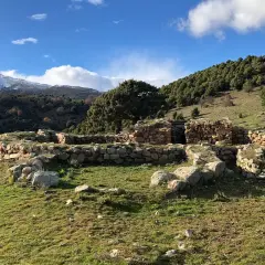 Antiche rovine di pietra, s'arcu e is forros, su un prato verde sotto un cielo azzurro con nuvole bianche e montagne innevate sullo sfondo.