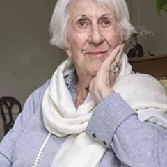 Annick de Souzenelle, con un sorriso sereno, appoggia il viso sulla mano, avvolta in una sciarpa chiara.