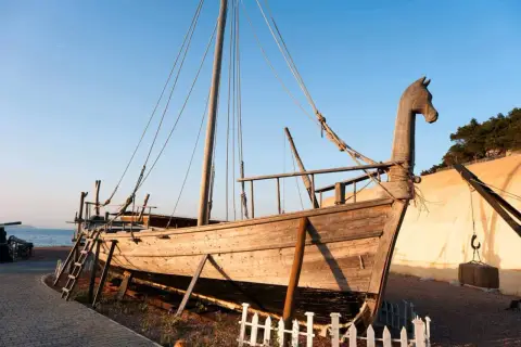 Una nave fenicia in legno, con prua a forma di testa di cavallo, è esposta su un terreno sabbioso vicino al mare.