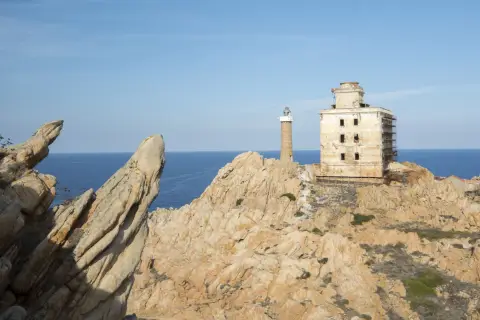 Il faro della Maddalena, con la sua torre e l'edificio adiacente in rovina, si erge su una costa rocciosa battuta dal mare.