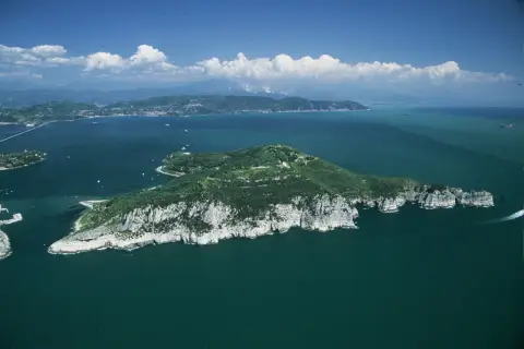 Isola verdeggiante con scogliere bianche, mare blu intenso e nuvole bianche. Si intravede la **grotta dei colombi** tra le rocce.