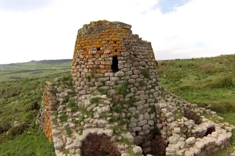 Il nuraghe Santa Barbara, con la sua torre in pietra ricoperta di licheni arancioni, domina un paesaggio verdeggiante e collinare.