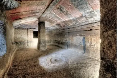 Interno di una tomba etrusca con soffitto dipinto e un pilastro centrale. Un cerchio inciso sul pavimento sembra un antico incantesimo putifigari.