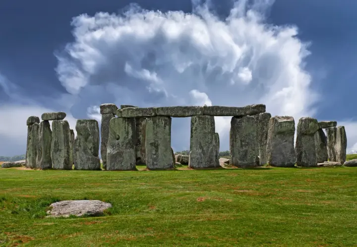 Antico cromlech di pietre imponenti su un prato verde, sotto un cielo drammatico con nuvole bianche e blu.