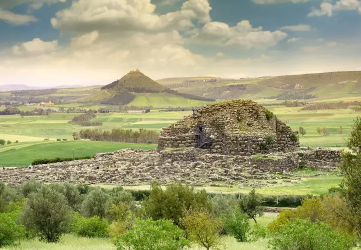Antico nuraghe in pietra, con scale esterne, immerso in un paesaggio verdeggiante con colline ondulate e un cielo nuvoloso.