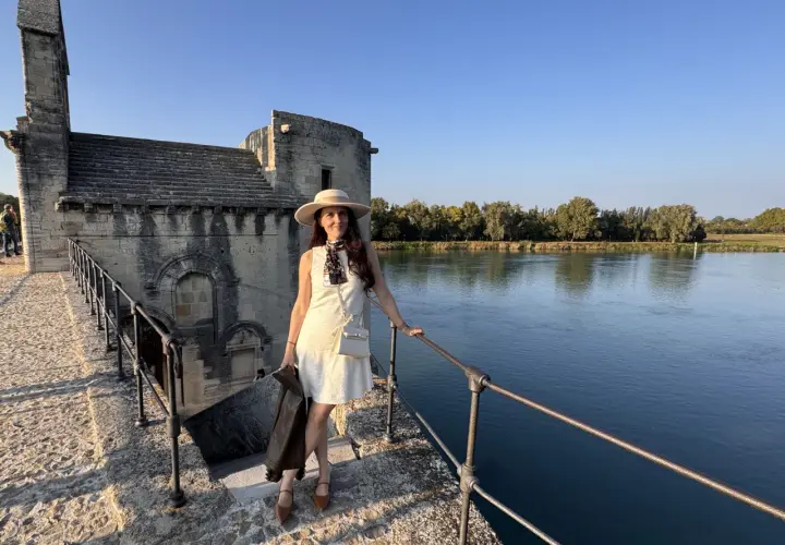 Donna con cappello e giacca in pelle, in piedi sul ponte di pietra vicino al fiume, con un edificio antico sullo sfondo, che ricorda un altare rupestre di Santo Stefano.