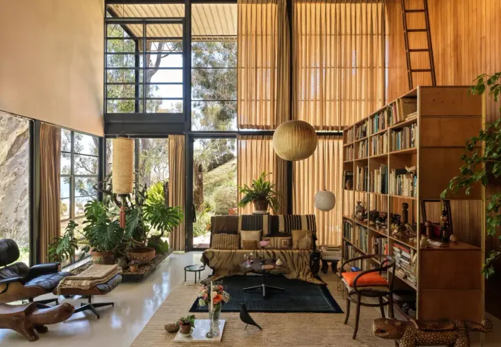 Interno della Eames House con grandi finestre, mobili iconici, piante rigogliose e una libreria piena.