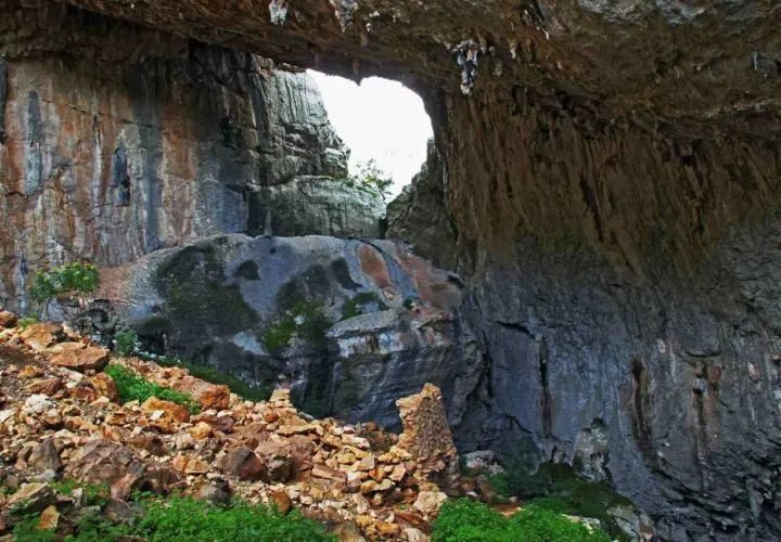 Ingresso di una grotta con rocce stratificate e vegetazione. Un arco naturale si apre verso l'esterno, rivelando un paesaggio luminoso. Un sito archeologico suggestivo.