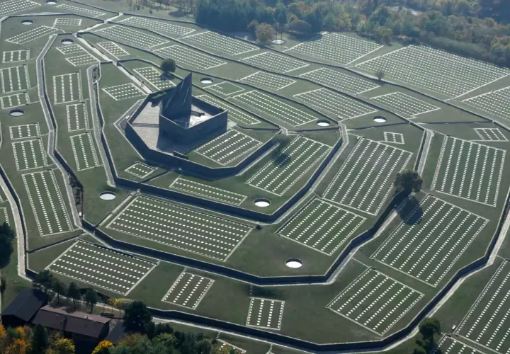 Vista aerea del cimitero tedesco al Passo della Futa, con file ordinate di croci bianche su prati verdi e un imponente monumento centrale.