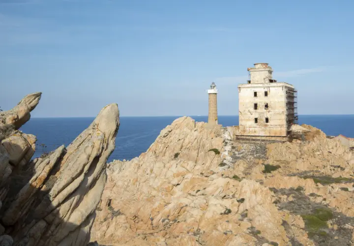 Il faro della Maddalena, con la sua torre e l'edificio adiacente in rovina, si erge su una costa rocciosa battuta dal mare.