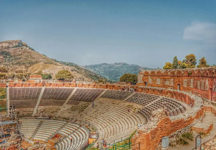 Antico teatro greco in Italia, con gradinate in pietra e rovine in mattoni rossi, incastonato tra colline verdi e un cielo azzurro.