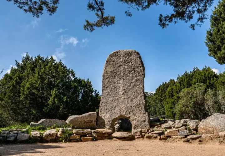 Un imponente menhir con un arco alla base, circondato da pietre antiche e vegetazione rigogliosa. Tra le tombe dei giganti più belle, questo sito archeologico incanta.