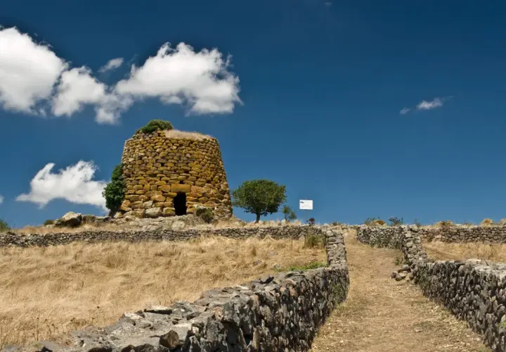Antichi nuraghi sardi, testimoni di un passato di guerra e civiltà. Un sentiero di pietra conduce verso la torre, immersa in un paesaggio arido sotto un cielo azzurro.