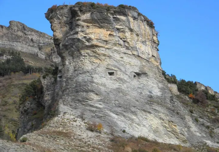 Imponente roccia con aperture che ricordano il vallo alpino del littorio. Un paesaggio montano suggestivo sotto un cielo azzurro.