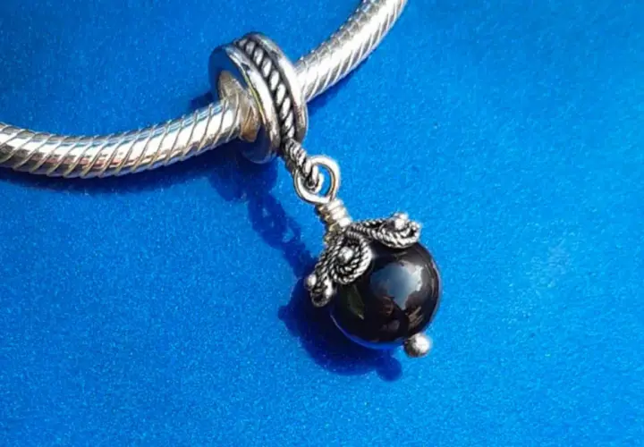 Un ciondolo con una pietra nera lucida, ispirato agli antichi amuleti sardi, pende da un bracciale d'argento su sfondo blu.