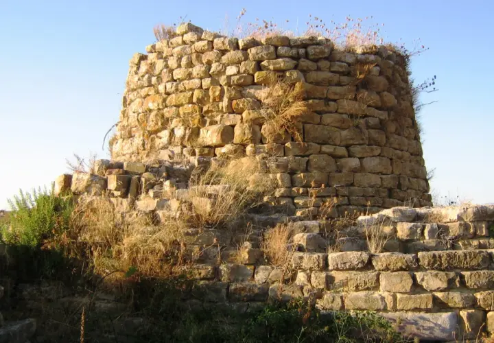 Il nuraghe Piscu, con le sue pietre antiche e l'erba secca, si staglia contro il cielo azzurro, testimone silenzioso di storie millenarie.