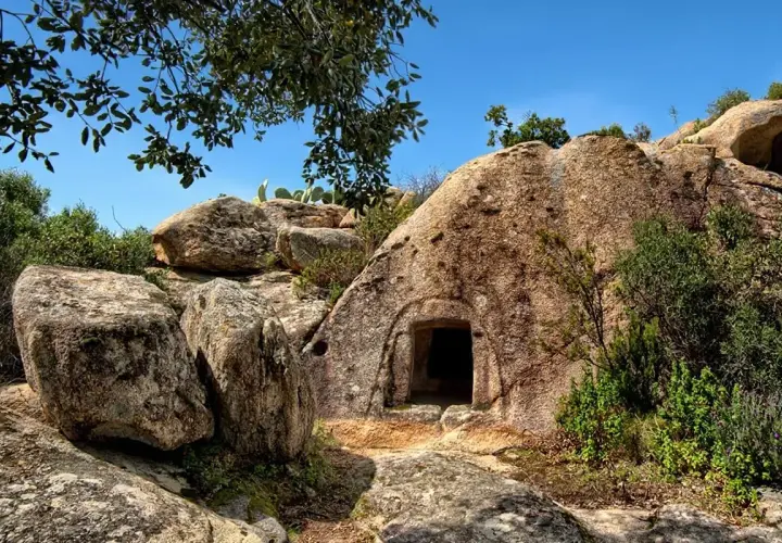 Ingresso di una delle **domus de janas**, tombe ipogeiche scavate nella roccia in Sardegna, circondata da rocce e vegetazione mediterranea.