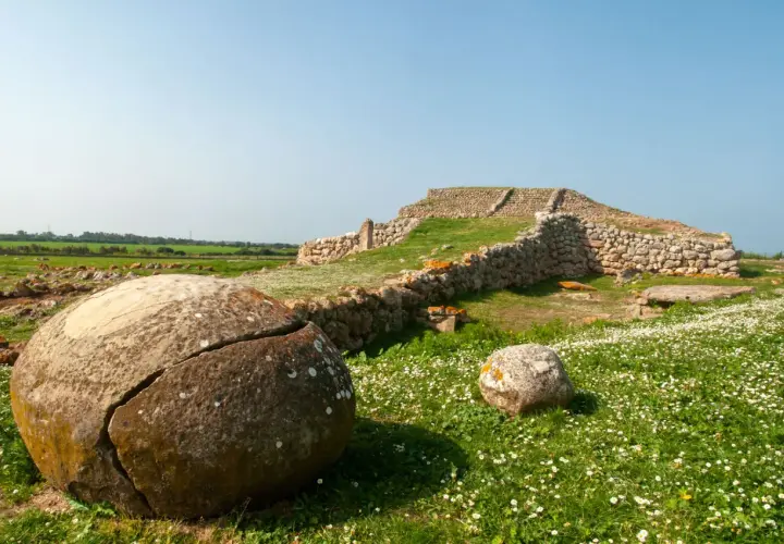 Antiche pietre e rovine di un ziqqurat in Sardegna, circondate da un prato fiorito e rocce tondeggianti.