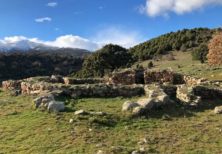 Antiche rovine di pietra, s'arcu e is forros, su un prato verde sotto un cielo azzurro con nuvole bianche e montagne innevate sullo sfondo.