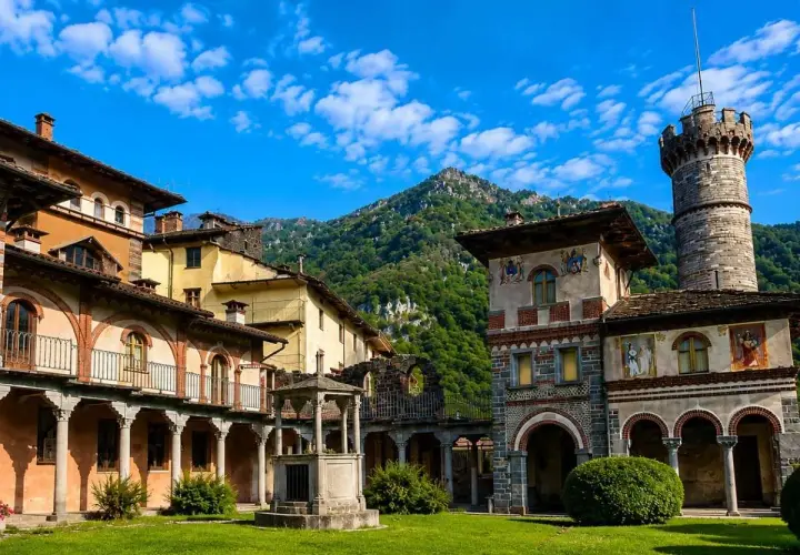 Cortile di un complesso architettonico con torre, affreschi e un pozzo, immerso nel verde. Sembra un luogo avvolto da un'aura di mistero, quasi una rosazza esoterica.