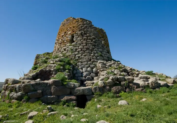 Il nuraghe Santa Barbara a Villanova Truschedu, imponente struttura in pietra con vegetazione rigogliosa, si erge sotto un cielo azzurro.