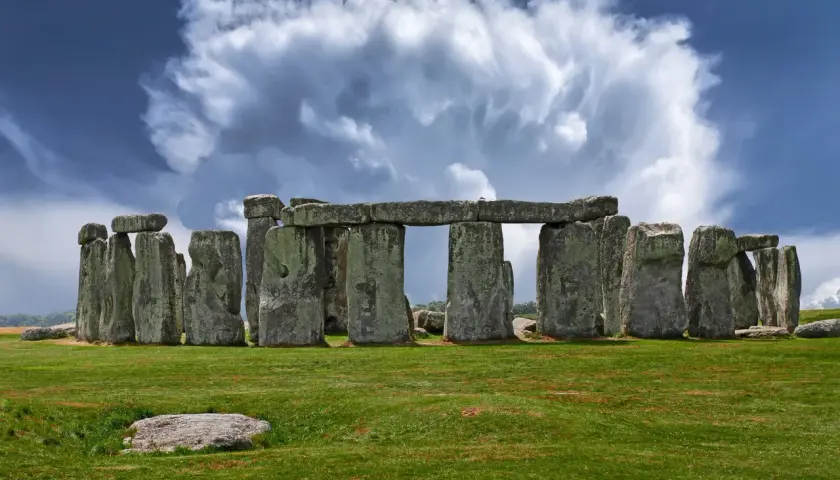 Antico cromlech di pietre imponenti su un prato verde, sotto un cielo drammatico con nuvole bianche e blu.