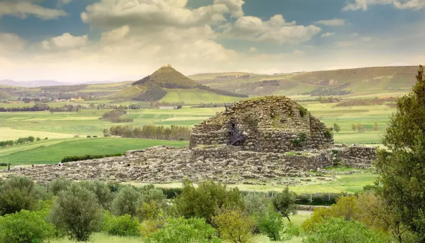 Antico nuraghe in pietra, con scale esterne, immerso in un paesaggio verdeggiante con colline ondulate e un cielo nuvoloso.