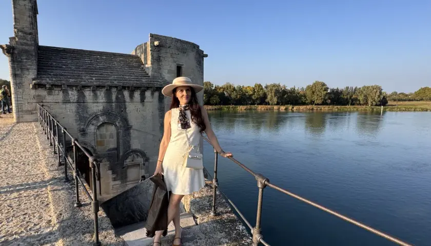 Donna con cappello e giacca in pelle, in piedi sul ponte di pietra vicino al fiume, con un edificio antico sullo sfondo, che ricorda un altare rupestre di Santo Stefano.