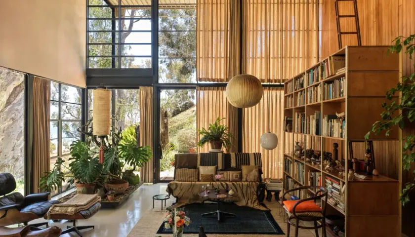 Interno della Eames House con grandi finestre, mobili iconici, piante rigogliose e una libreria piena.