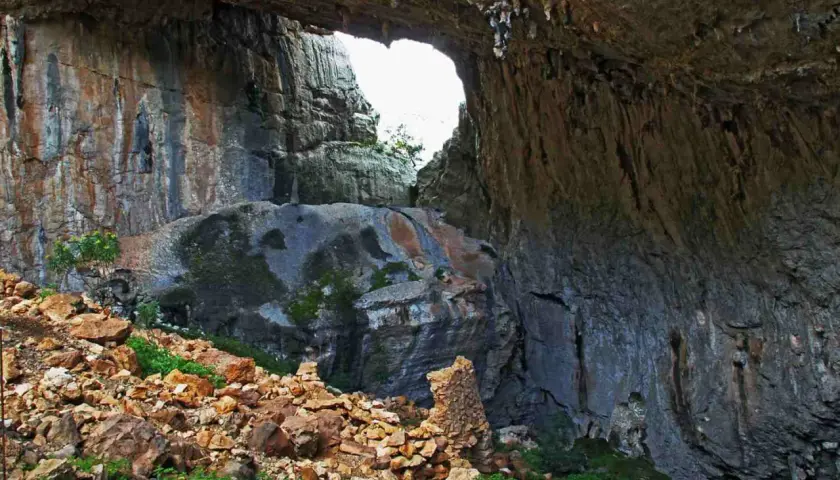 Ingresso di una grotta con rocce stratificate e vegetazione. Un arco naturale si apre verso l'esterno, rivelando un paesaggio luminoso. Un sito archeologico suggestivo.