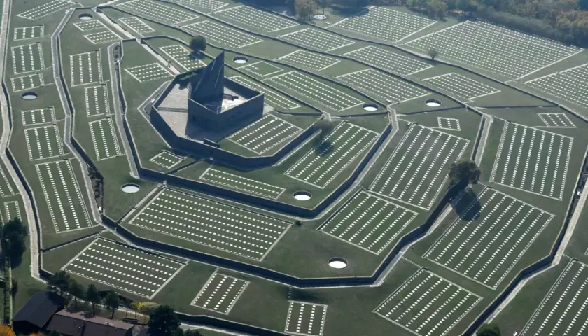Vista aerea del cimitero tedesco al Passo della Futa, con file ordinate di croci bianche su prati verdi e un imponente monumento centrale.