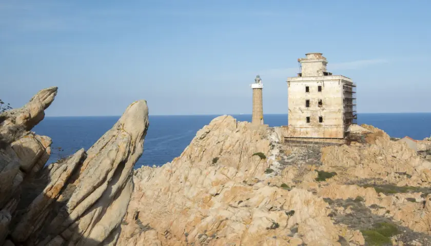Il faro della Maddalena, con la sua torre e l'edificio adiacente in rovina, si erge su una costa rocciosa battuta dal mare.